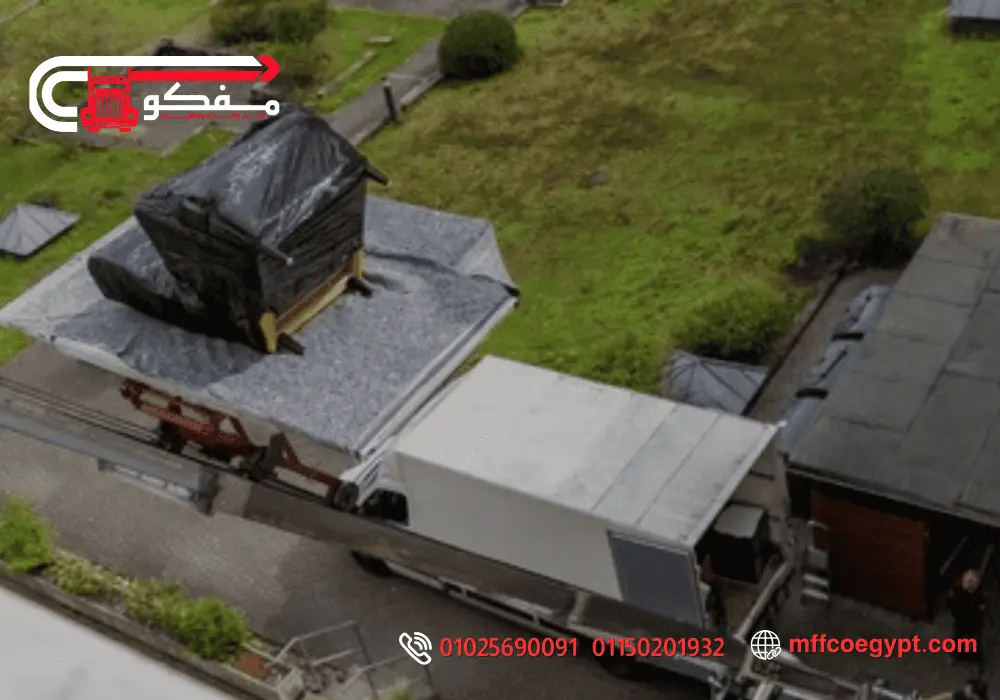 ارخص ونش رفع الاثاث في مصر الجديدة - نقل آمن وسريع للأثاث الكبير A truck with a flatbed carrying a large item covered in black protective material on a gravel surface, driving on a street near grass and buildings.